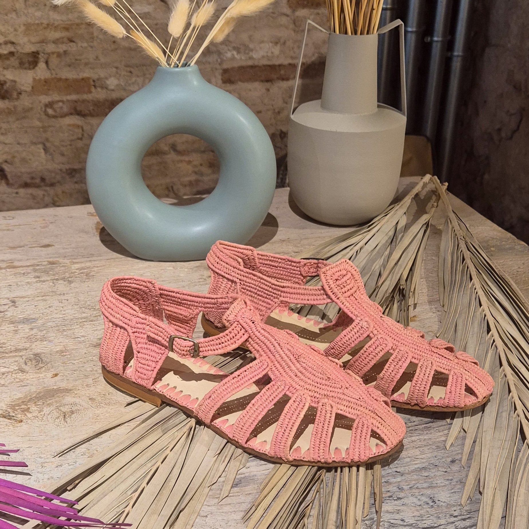 Cangrejera Hana en tono rosa empolvado sobre hojas de palmera secas, cerca de un jarrón redondo con un agujero en el centro en tono azul pastel