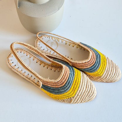 Sandalia Amel de Leke's de rafia natural, un diseño slingback sobre un mueble blanco junto a un jarrón gris.