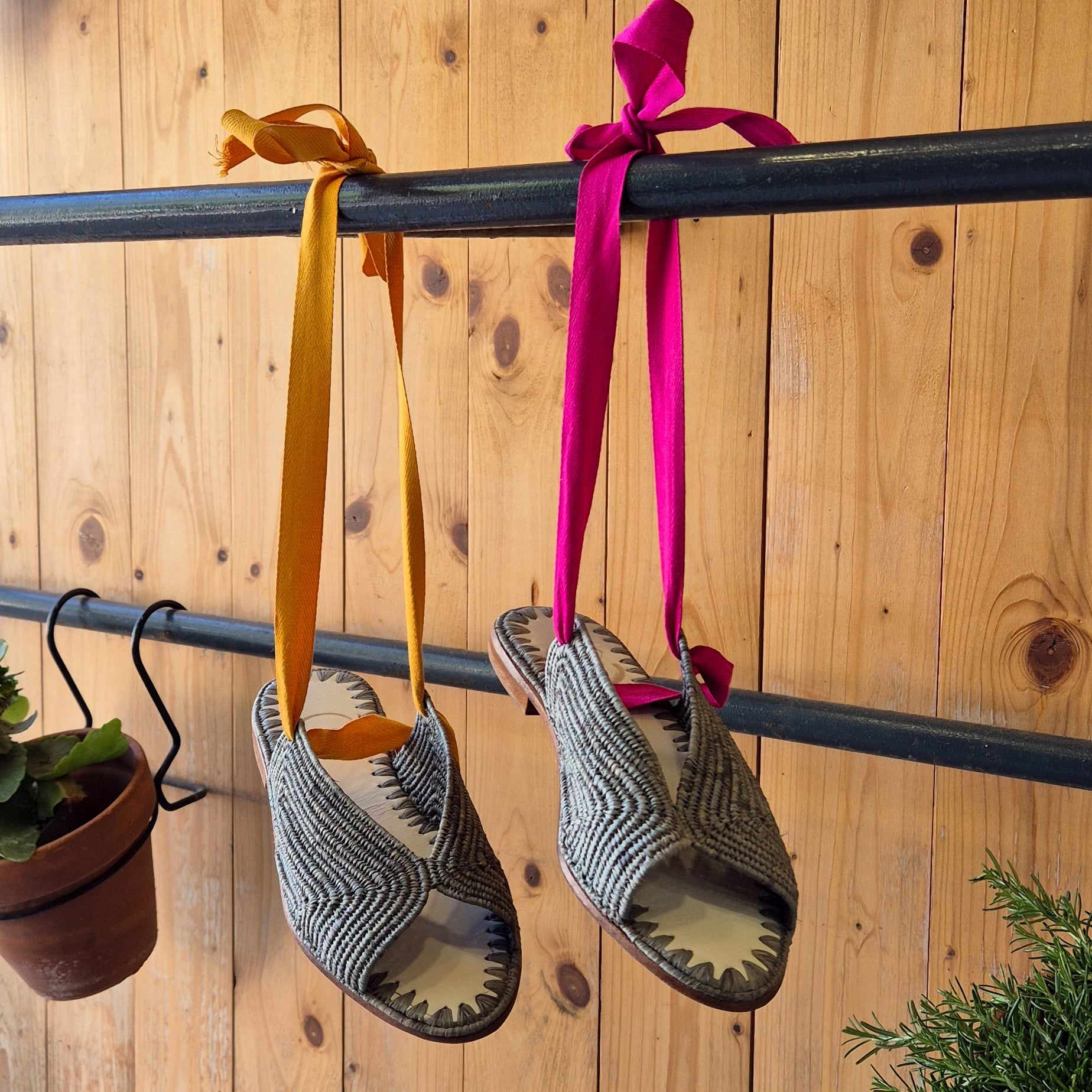 Sandalias Fara en tono gris con cintas de algodón en tono mostaza y fucsia colgadas de una barra de metal