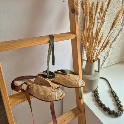 Sandalia Fara de Leke's en tono rafia natural con las cintas de algodón en marrón y en verde aceituna sobre una escalera de madera de pino 