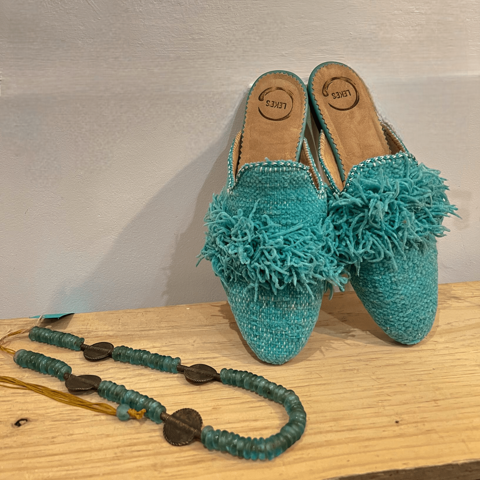 Mule Salma de Leke's apoyado en pared blanca encima de un banco de madera natural y al lado un collar de piedras naturales a conjunto con el mule 